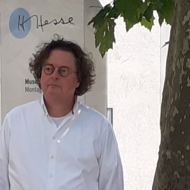 Mann mit Brille in weißem Hemd, steht vor einer Wand mit Logo und Baum.